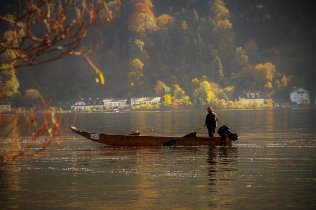 A fisherman is stood in a wooden rowing boat in a large body of water. In the background there are houses along the waterfront and trees covering the side of the valley behind them. 