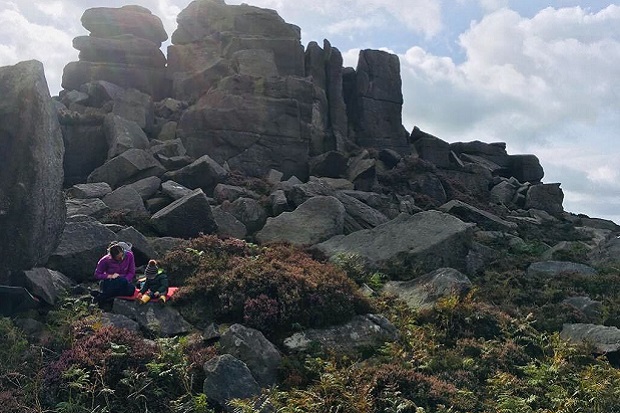 A sunny day in the Peak District. There are tall rocks in the background with lots of boulders, moss and ferns in front of them. Jeni and a toddler are sat on a blanket on a rock near the front, wearing coats and hats.  