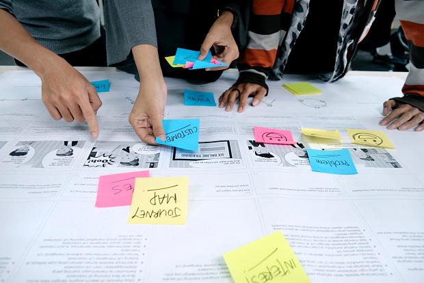 A large format print is covering a table with text and images. Three people are leaning over the table, sticking colourful post-it notes in different places. The post it notes have writing on, such as ‘journey map’, and ‘problem’. 