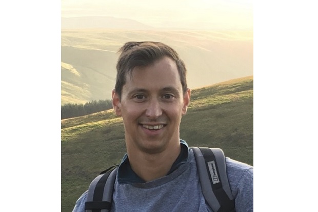 Jan is stood high up on a hillside, smiling and wearing a jumper and rucksack. The sun is glowing on the fields and hills behind him. 