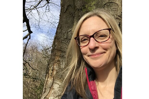 Laura is wearing glasses and smiling at the camera. She’s stood in front of a large tree trunk, with more trees and blue sky behind her. 