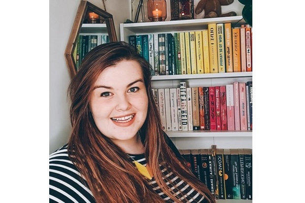 Alice sits smiling in front of a bookcase full of colourful books. She is wearing a top with black and white horizontal stripes and her long brown hair is worn down and parted to the side. 