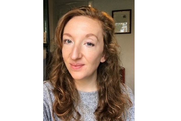 A lady, with long wavy, auburn hair, wearing a grey jumper, smiles at the camera.