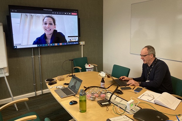 On the right of the picture, A man on a laptop, sitting at a desk with other compute equipment visible and a water bottle. On the left, a lady appears on a large screen, smiling.