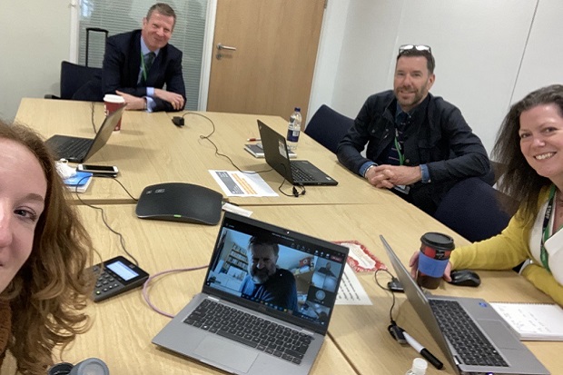 Four people in an office, two men and two women, with laptops on the desk and some with drinks,, all looking at the camera.