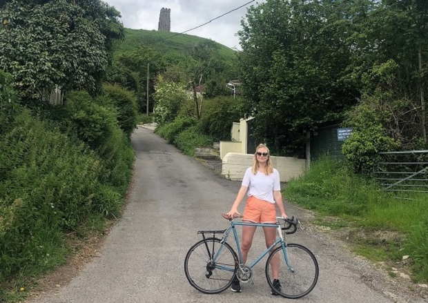 Anna is stood holding her bicycle on a single track road. She is wearing sunglasses, a white top and orange shorts. Above her you can see Glastonbury Tor.