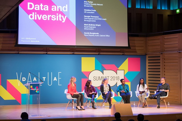 Anna is sat to the left of a panel of experts at the Open Data Institute annual summit in 2018. She wears a bright orange shirt and black trousers, with a radio mic on her lapel. There is a big sign behind them saying ‘Data and diversity’