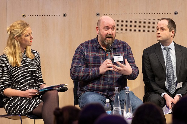 Anna is sat to the left of two people on a panel. She is wearing a black and white dress and holding a microphone. 