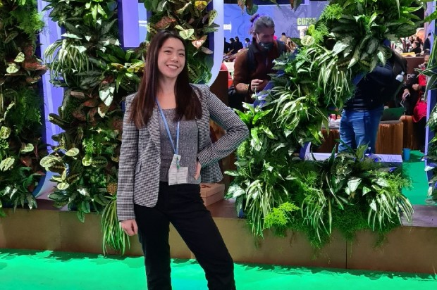 Emily is smiling, standing in front of a leafy display at a work conference. She is wearing a grey blazer and black trousers, with a lanyard round her neck.