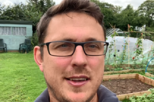 A man, with a beard, dark hair and glasses, standing on a allotment, with a shed in the left of the background and a polytunnel to the right of him.
