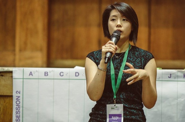 Emily is speaking at a work event. She is holding her microphone in her right hand and her left hand is gesturing. She is wearing a black top with a green lanyard round her neck.