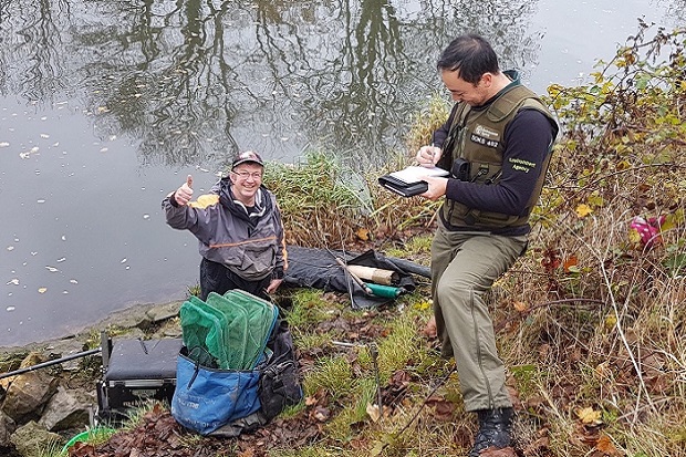 Alt text: An Environment Agency Fisheries Enforcement Officer holds a pad in one hand as he checks a licence while an angler smiles on the bankside.