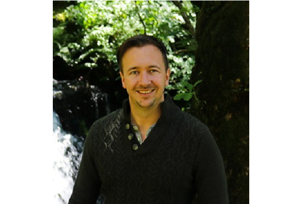 A white man, with short hair and wearing a dark sweater, in front of tress and foliage, with water cascading in the background too.