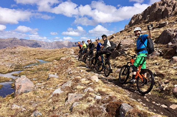 A group of cyclists are lined up on a path in the Scottish Highlands. They are looking back and smiling at the camera. Behind them is a blue sky with some small, white clouds.