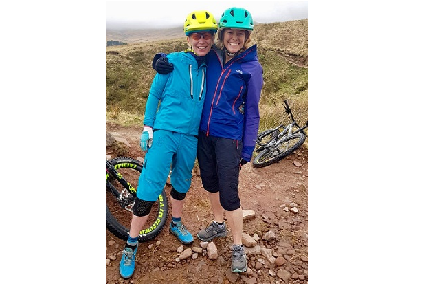 Steph stands with BBC presenter Kate Humble in the Brecon Beacons. Their bikes are behind them on the ground. Kate wears a dark blue waterproof jacket and a lighter blue helmet. She has her arm around Steph. Steph wears a light blue waterproof jacket and a yellow helmet.