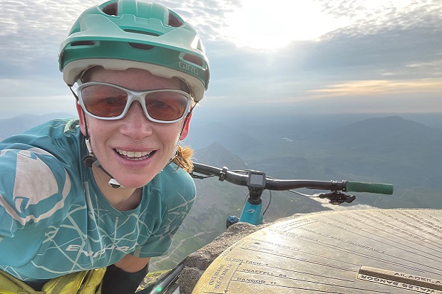 Steph with her bike on the top of Snowdon, a mountain in Wales. Behind her, the sun is shining through clouds. Steph is wearing sunglasses, and a blue cycling helmet with a matching top.