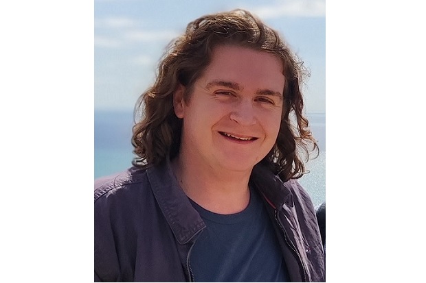 A man with shoulder length wavy hair, wearing a blue t shirt and zip top, smiles at the camera. Blue sky is in the background.