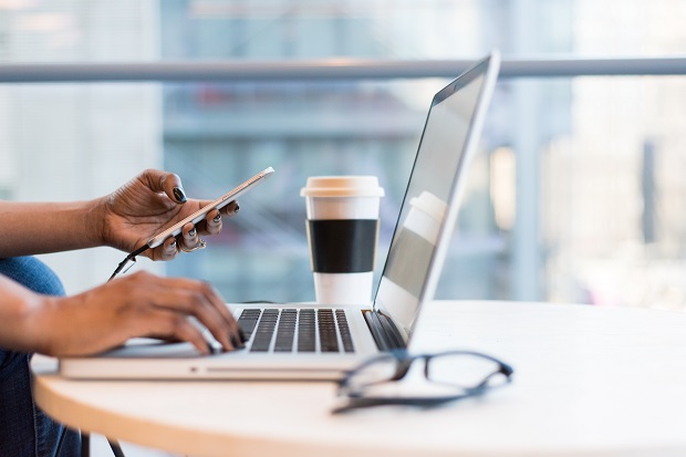 A person's hands, one hand typing on a laptop keyboard and the other hand holding a mobile phone, with a pair spectacles in the foreground and a coffee cup in the background.