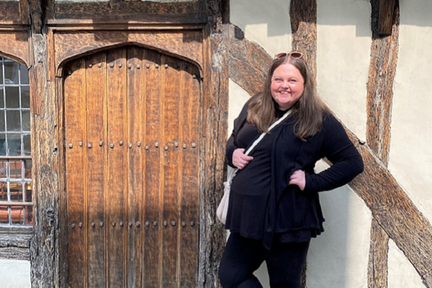 A lady, smiling, with long hair, a white bag over her shoulder, sunglasses on head and dressed in black, stands next to the carved wooden door of an old building.