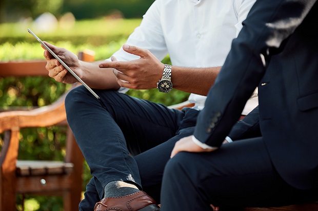 A picture containing two people, sitting outdoors, one holding a mobile device while showing the other person something on the screen.