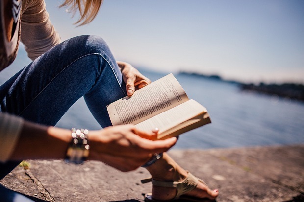 A lady, wearing jeans, sandals, and a bracelet, reading a book by the water.