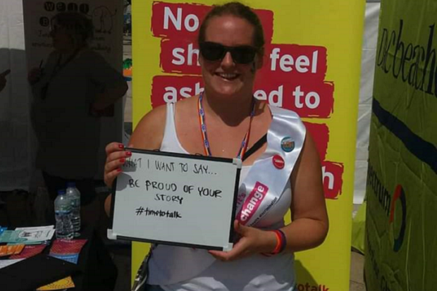 A lady, standing in front of a yellow and red poster, wearing a white vest and sunglasses, and holding up a sign that says ‘What I want to say, be proud of your story, hashtag time to talk’.