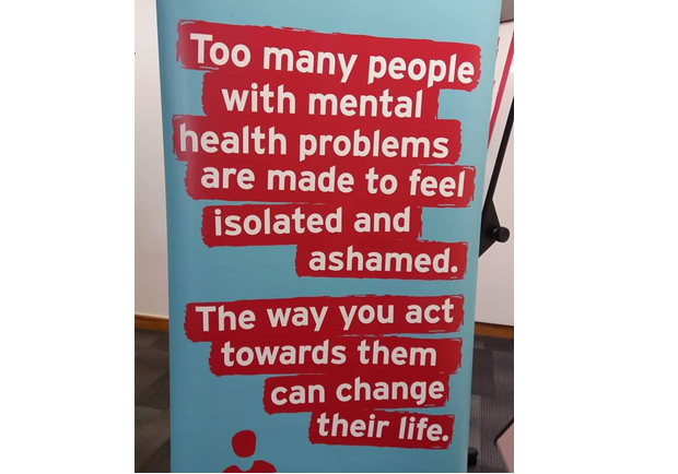 A sign, in white writing on a blue and red background, which says ‘too many people with mental health problems are made to feel isolated and ashamed, the way you act towards them can change their life’.