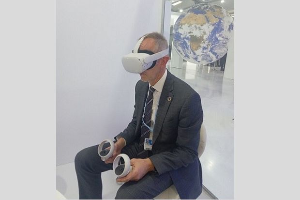 A man in a grey suit and striped tie, wearing a virtual reality headset and using virtual reality hand paddles.