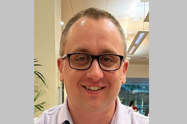 A man with short hair, black glasses, and wearing a light-coloured plain shirt., with a window in the background to the right, and a plant partially visible to the left.