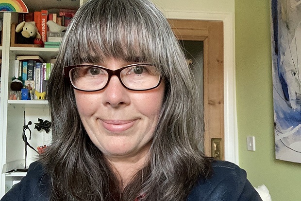 A lady, smiling, with long hair, glasses and blue shirt, a bookcase and books in the left of the background and a picture to the right.