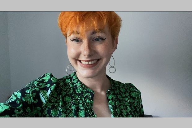 A lady, smiling, with short orange hair, large earrings, and a green and black patterned blouse.