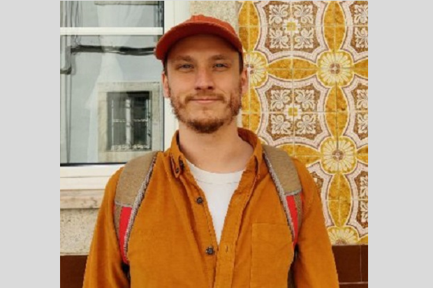 A bearded man wearing a red cap, brown jacket and with a backpack, a window in the background to the left and yellow patterned wallpaper to the right.