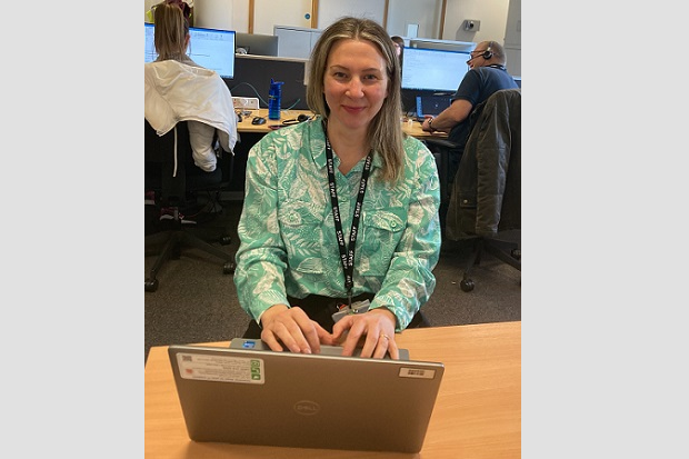 A lady, smiling, with long straight hair and wearing a headset, sits in front of a laptop at a desk.