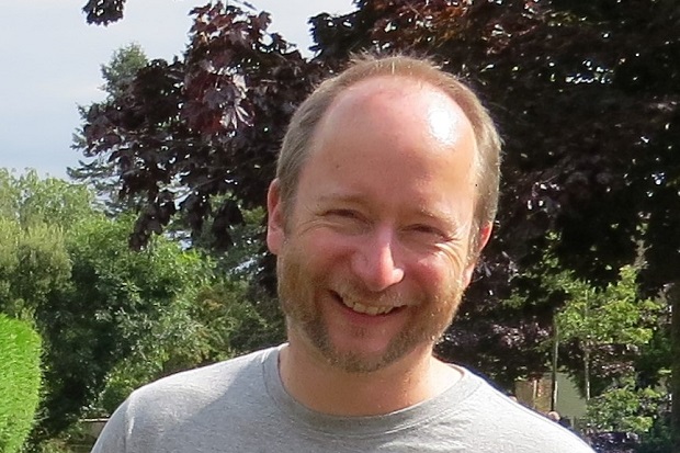 A man with short, receding hair, and a beard with trees and bushes in the background.