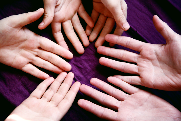 The palms of six different hands, in a circle, on a purple cloth backdrop