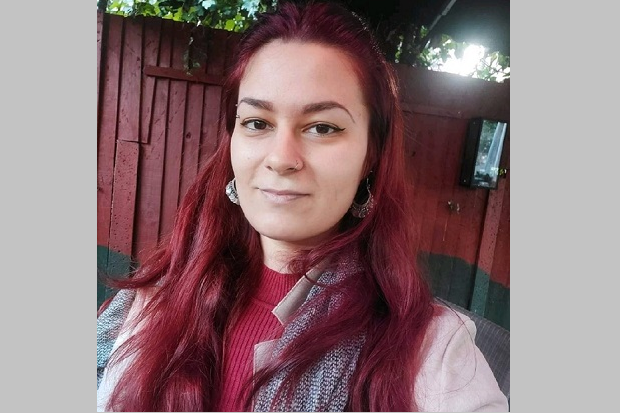 A lady, sits, smiling, with long pinkish hair, silver earrings, wearing a red sweater and grey jacket.