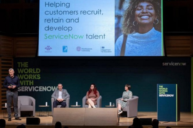 Four people on a stage (two men, two women), the man on the left is standing, the others are all sitting, with a sign behind them which says 'helping customers recruit, retain and develop Service Now talent.