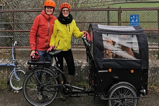 Two ladies, dressed in cycling clothing, stand in front of a bicycle with a dog basket attached to the front.