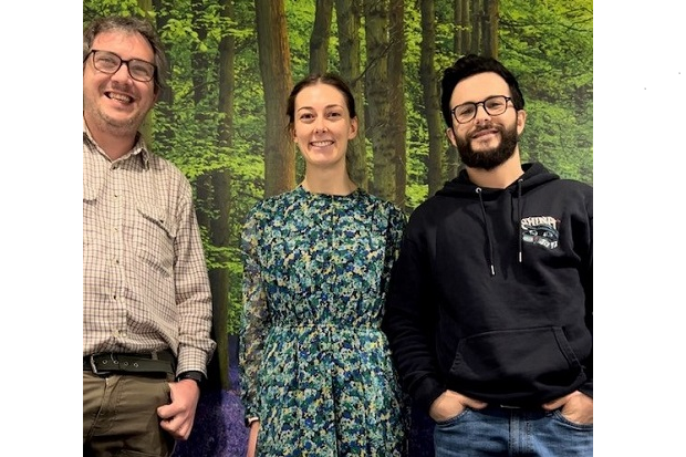 On the left, a man with curly hair, glasses, beige shirt and brown trousers, centre, a woman in a blue and green floral dress with hair tied back, and right, a man with short dark hair, glasses and beard, wearing a blue hoodie and jeans.