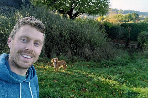 A man, in a field, wearing a blue hoodie, with a brown dog in the background and trees.