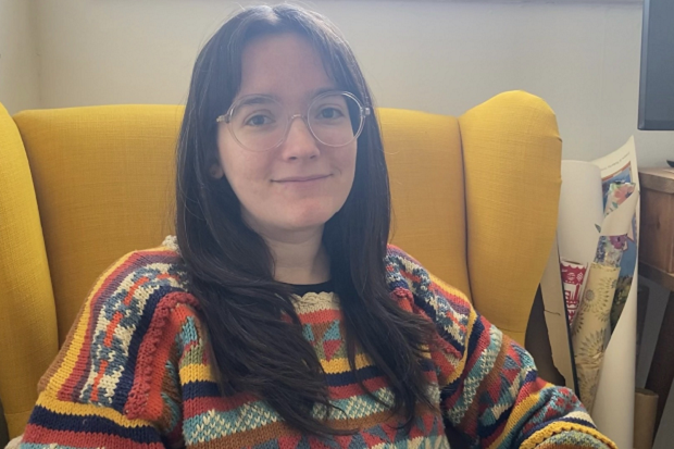 A lady with long, dark brown hair, wearing a stripy jumper and sitting in a yellow armchair.