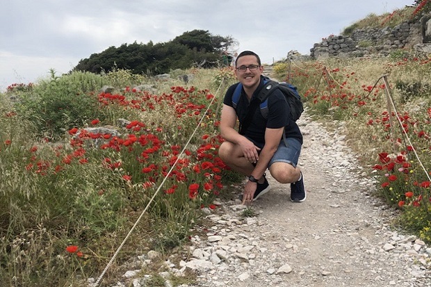 A man in shorts, t shirt and a backpack, wearing glasses, kneeling down on a path in font of some red flowers.