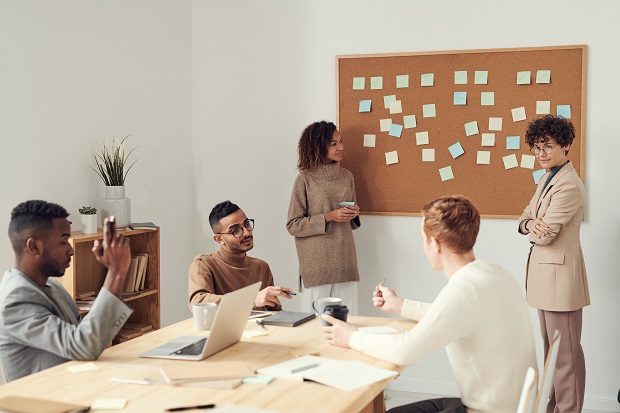 A group of people in a meeting, sat around a table, and post it notes stuck onto a cork board that is hanging on the wall.