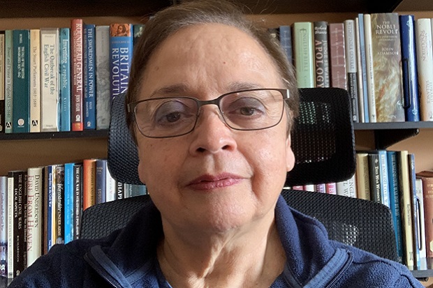 A lady with short hair and glasses, a selection of books on shelves in the background.