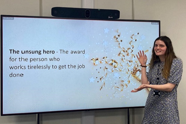 A lady, with long dark hair and wearing a floral print dress, stands and gestures to a large TV screen to her right which displays the words 'the unsung hero - the award for the person who works tirelessly to get the job done'.
