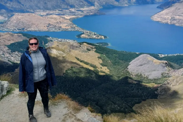 A lady wearing sunglasses, a grey zip top and blue coat, stands on a hill with a blue lake in the background.