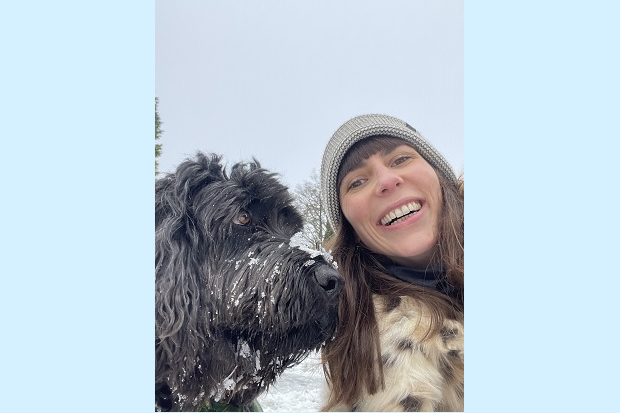 On the left, a large black dog with snow on their nose, on the right a lady, with long dark hair and a hat, smiling.