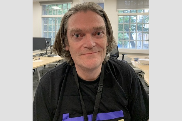 A man, with shoulder length brown hair, wearing a black t shirt and lanyard, with desks, a computer and windows in the background.