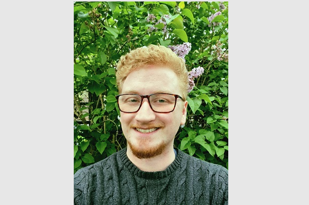 A man with short ginger hair and short ginger beard, dark glasses and a grey sweater.