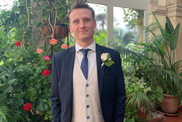 A man with short brown hair wearing a blue suit and tie, with cream waistcoat and a white rose on the lapel.